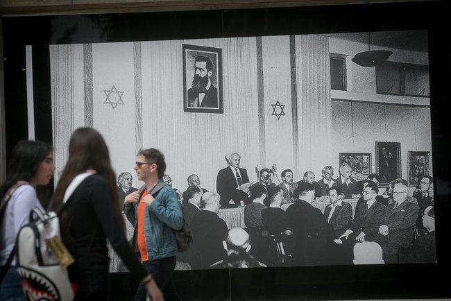 Люди проходят мимо большой фотографии, изображающей Декларацию государства Израиль, написанную первым премьер-министром Давидом Бен-Гурионом