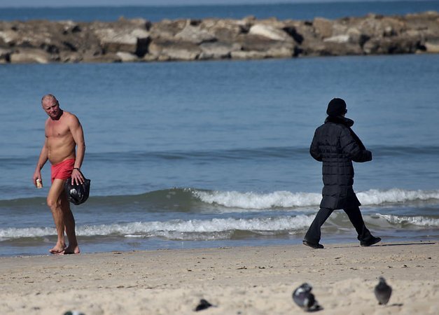 Мужчина в плавках проходит мимо женщины в зимней одежде на пляже в Тель-Авиве
